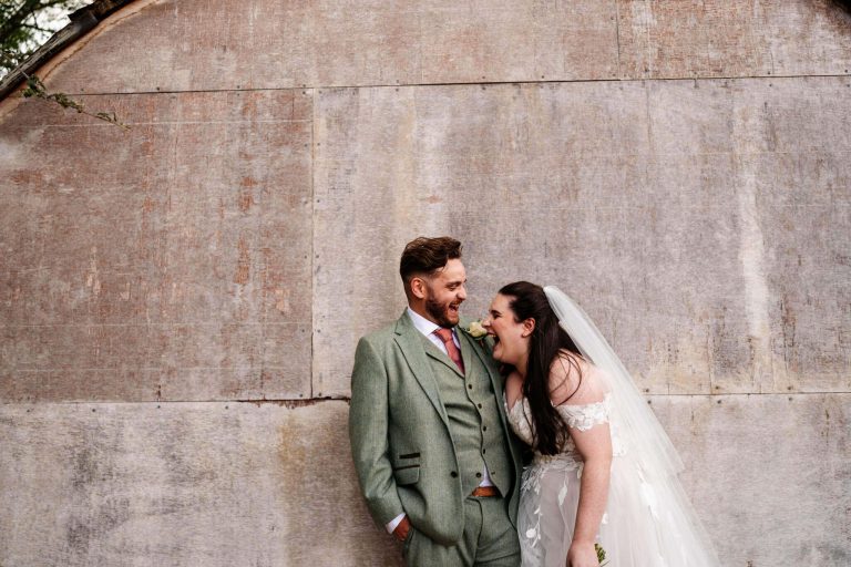 Bride and groom share a funny joke whilst standing next to a concrete wall at Carriage Hall Nottingham