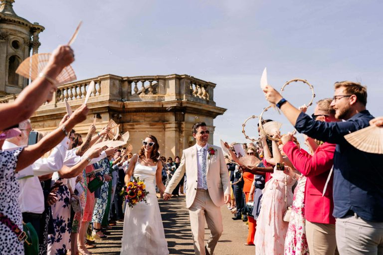Bride and groom walk holding hands between rows of their friends and family waving fans in place of confetti with Normanton Church in the background after their wedding ceremony