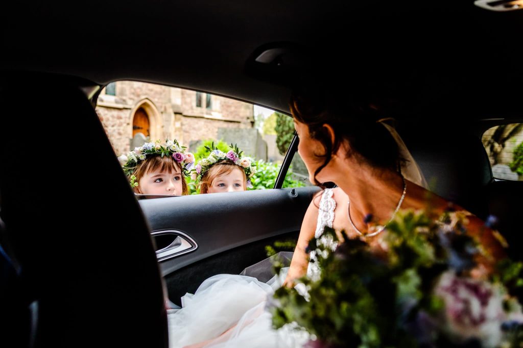A bride in a car looks at two young girls outside, who are wearing flower crowns and peering in through the window. The background shows a stone building and greenery. The focus is on the bride and the girls interaction.