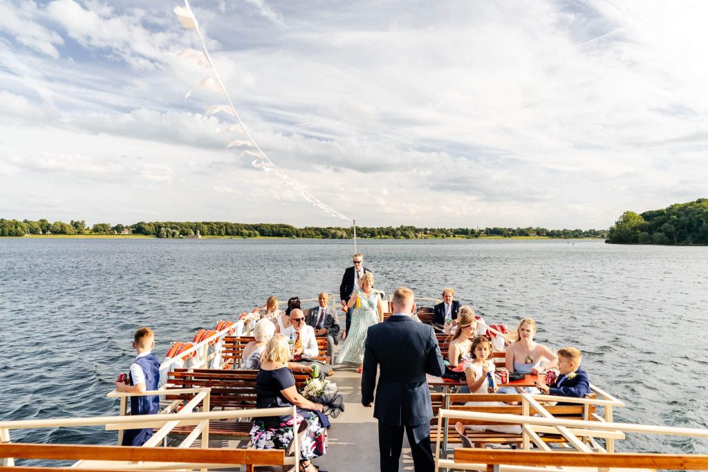 A group of well-dressed people are gathered on a boat on Rutland Water, against a scenic lake background with lush green trees and a partly cloudy sky. They appear to be in conversation, enjoying a pleasant day aboard the Rutland Belle near Normanton Church.