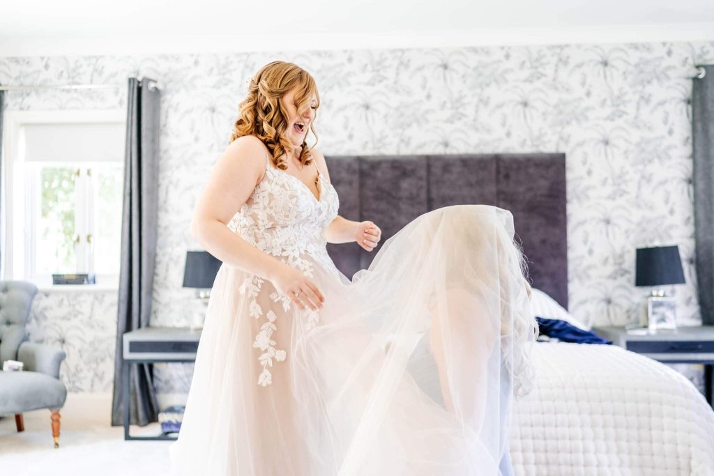 A bride in a white lace gown stands in a bedroom with elegant wallpaper. She laughs joyfully as her sister gets stuck in her dress whilst trying to help her