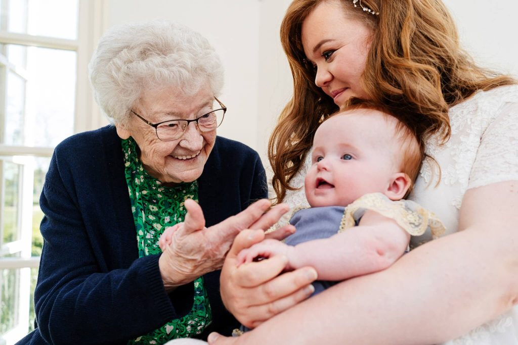 An elderly woman with glasses and a green blouse smiles warmly at a baby. A woman with long hair holds the baby gently in her arms. They are sitting together in a bright room with large windows.