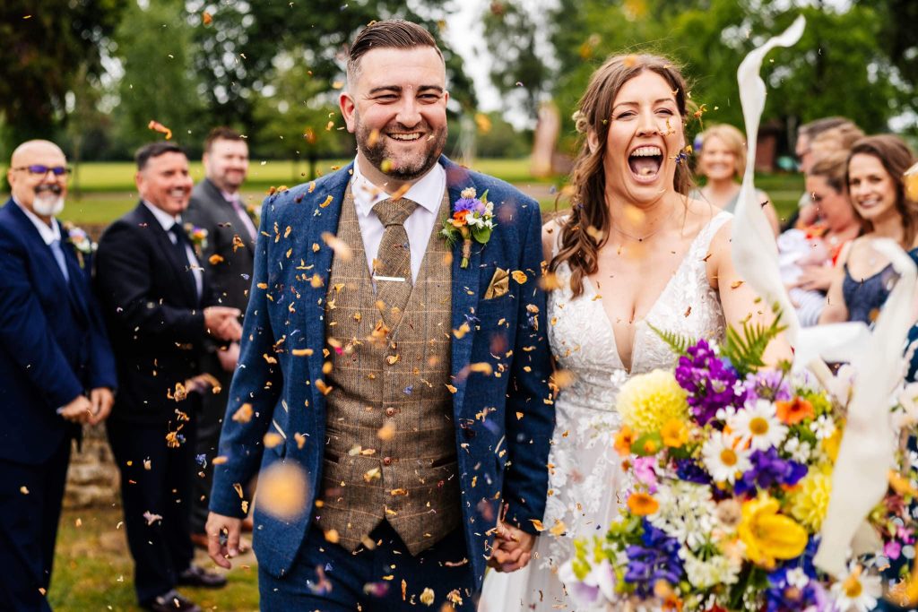 A joyful couple holding hands walks down an outdoor aisle, smiling broadly. They're surrounded by guests tossing colorful confetti. The bride holds a vibrant bouquet of flowers, and the groom wears a blue suit with a checkered vest.