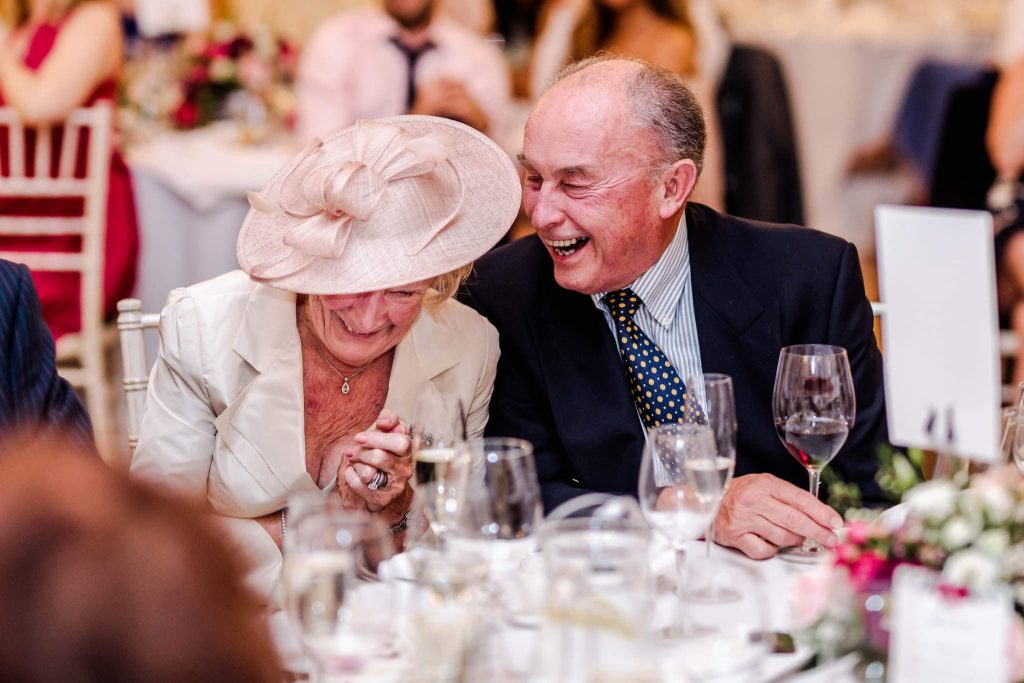 An elderly couple, elegantly dressed, share a joyful moment at a formal event held at the Old Naval College in Greenwich, London. The woman wears a stylish hat, and the man is in a suit and tie. They are seated at a table with wine glasses and floral decorations, smiling and laughing together.