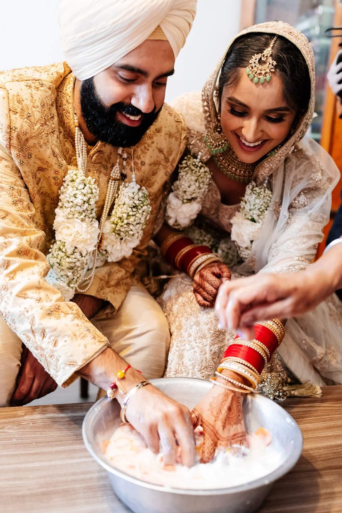 A smiling bride and groom, both in traditional attire, participate in a Sikh wedding tradition. They are dipping their hands into a bowl of milk and flower petals, searching for their ring. The bride wears henna on her hands, and the groom is in a cream turban.