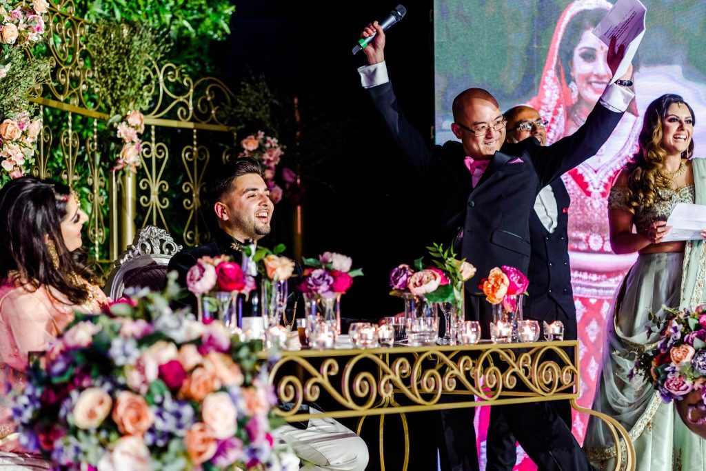 At a floral-decorated Hindu wedding reception at Kelham Hall in Nottinghamshire, a man in a suit joyfully raises a microphone while giving a speech. The couple, seated at their table adorned with colorful flowers and candles, smiles warmly as a large photo of the bride is displayed in the background.
