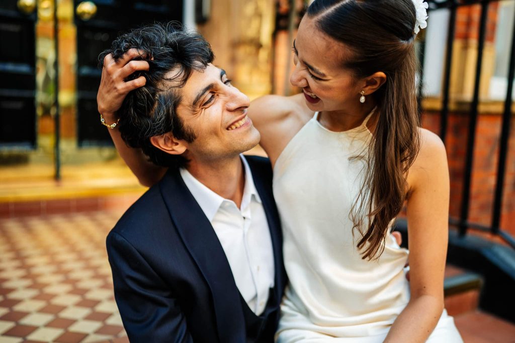 A joyful couple, elegantly dressed, share a loving moment after their Marylebone Town Hall wedding. The woman smiles as she sits on the mans lap, gently touching his hair. They are on a patterned floor with a door in the background, capturing the bliss of their special day.