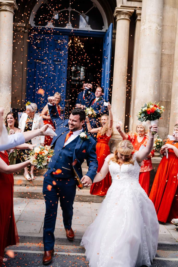 A joyful bride and groom celebrate outside Louth Town Halls large blue doors. Guests in red dresses and suits cheer and toss confetti. The bride clutches her bouquet, while the groom raises his hand in excitement.