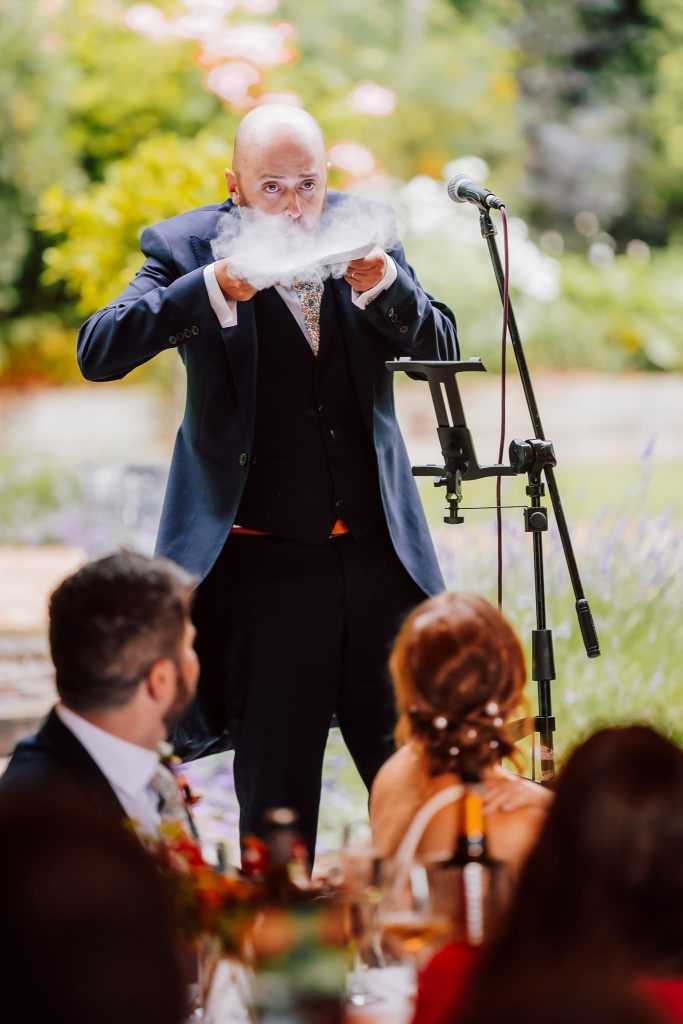 At Ashley Wood Farm in Wiltshire, a person in a suit dazzles an audience, conjuring mist from a bowl before a microphone. Guests are captivated by the magical display at this charming outdoor event, surrounded by lush greenery and blossoming flowers.