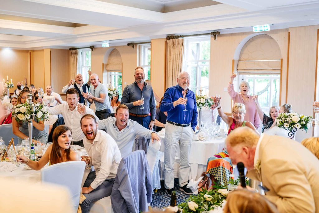 A lively wedding reception at Ardencote, with guests joyfully raising glasses and laughing. Tables are adorned with flowers, and people are seated in formal attire. The atmosphere is vibrant and festive, with natural light streaming through the windows.