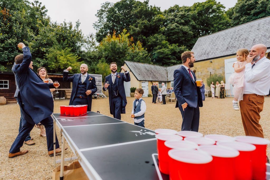 At Ashley Wood Farm in Wiltshire, a group of men in suits play beer pong outdoors, with one tossing a ball. A young boy in a vest observes keenly. In the background, a man cradles a child while others linger near the rustic buildings and trees. Red cups dot the table, infusing the lively scene with fun.
