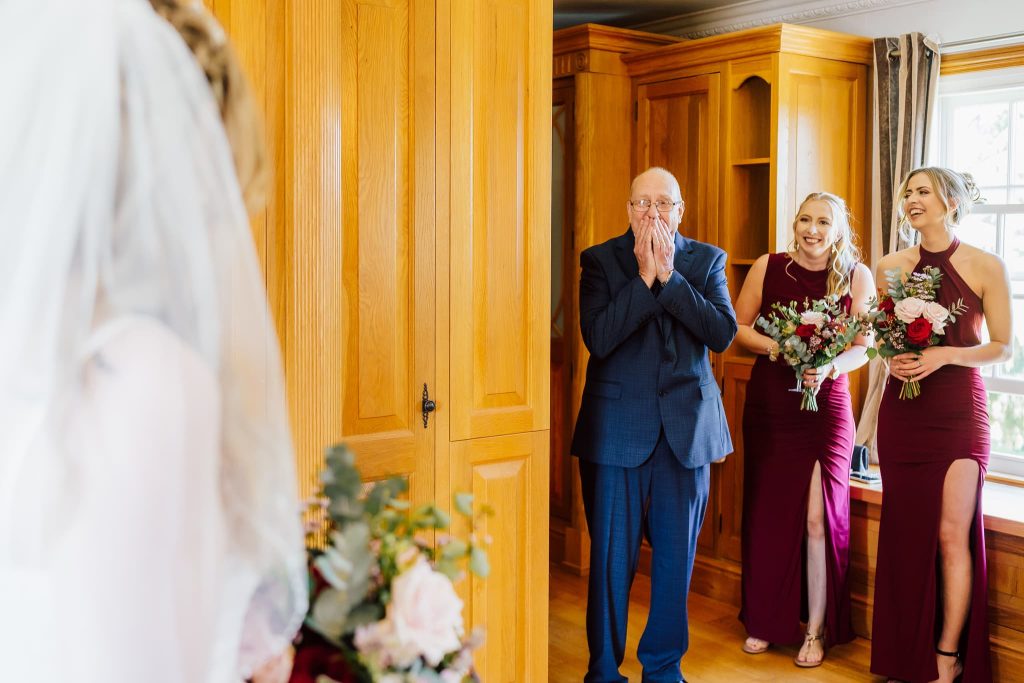 A bride in a white gown faces her father and bridesmaids. The father, wearing a blue suit, looks surprised and emotional, hands covering his mouth. The two bridesmaids in burgundy dresses hold bouquets and smile with joy. The setting is a warmly lit room.