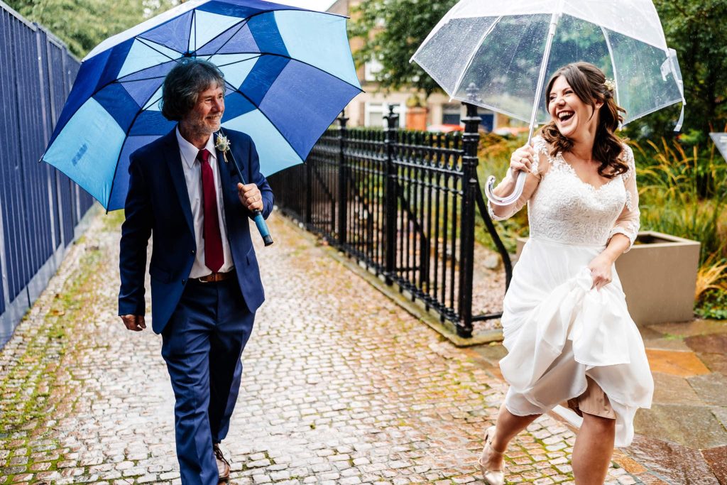 A joyful bride in a white dress playfully holds up her skirt while walking with a transparent umbrella. Beside her, a man in a suit with a blue umbrella smiles. Theyre on a cobblestone path with greenery in the background.