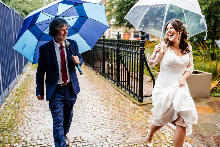A joyful bride in a white dress playfully holds up her skirt while walking with a transparent umbrella. Beside her, a man in a suit with a blue umbrella smiles. Theyre on a cobblestone path with greenery in the background.