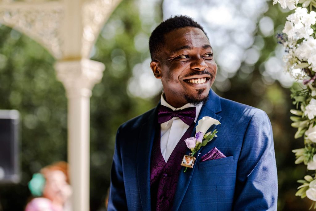 A man in a blue suit and bow tie smiles warmly while standing outdoors. He wears a boutonniere with a lily and purple flower on his suit lapel. The background is blurred, with greenery and flowers visible.