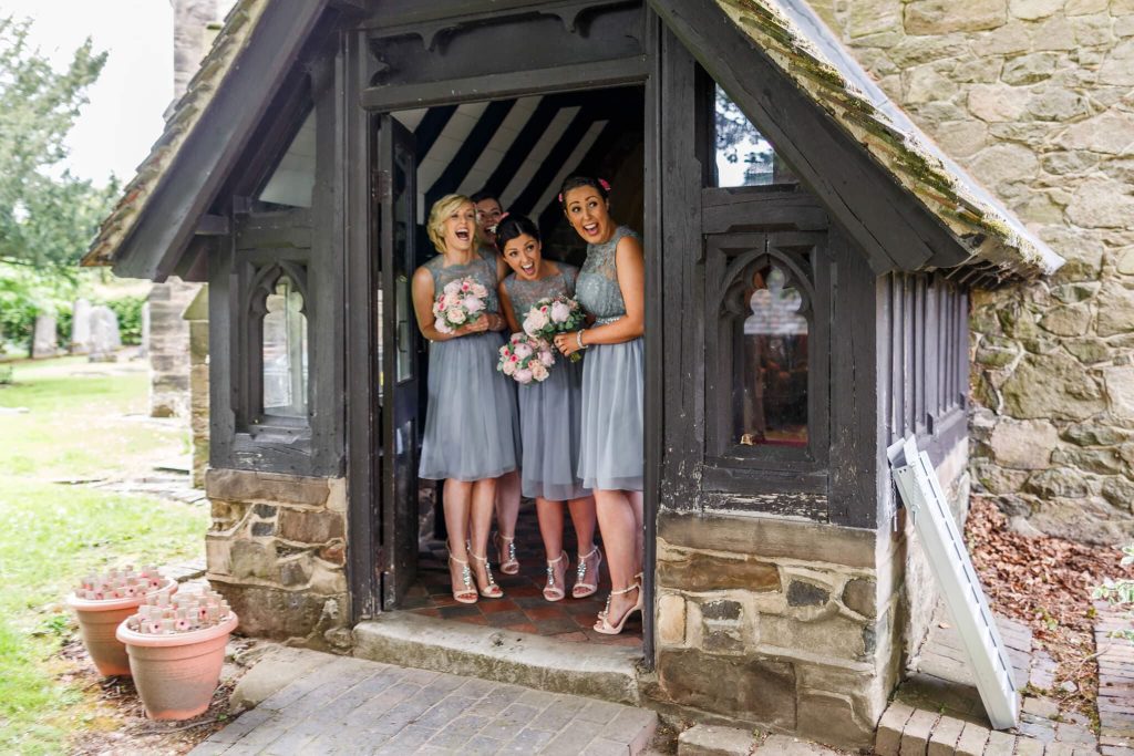 Four women in matching grey dresses stand in the entrance of a small, rustic chapel, holding bouquets of pink flowers. They are smiling and laughing, with stones and greenery visible outside.