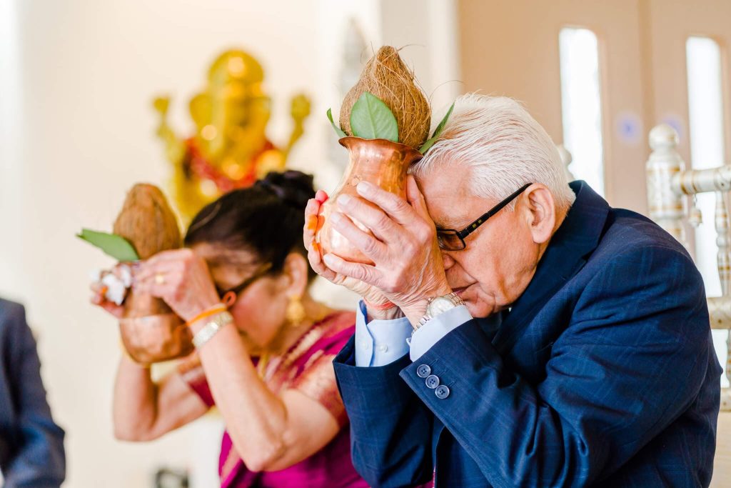 An elderly couple participates in a traditional Hindu ceremony, holding copper pots with coconuts and leaves against their foreheads. The man wears a suit, and the woman is dressed in a saree. A blurred religious statue is visible in the background.