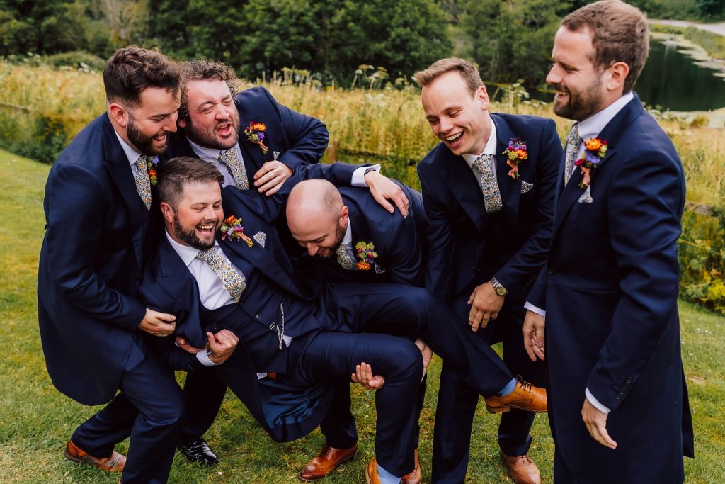 A group of six men in suits are laughing and playfully lifting one of their friends off the ground. They are standing on a grassy area with a lake and trees in the background. The mood is joyful and celebratory.