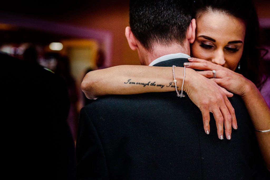 A couple embraces closely during a dance. The womans arm is wrapped around the mans shoulders, revealing a tattoo that reads I am enough the way I am. She is wearing a ring and a bracelet. The background is softly blurred.