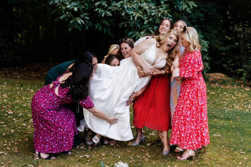 A joyful group of women in colourful dresses laugh and gather to lift a bride in a white gown. They are outdoors, surrounded by greenery and fallen leaves, creating a lively and playful scene.