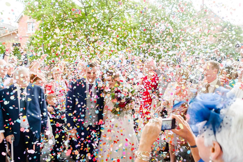 A newlywed couple walks through a colorful shower of confetti. The bride holds a bouquet, and the groom wears a suit. Guests surround them, some taking photos. Bright, sunny day with trees in the background.