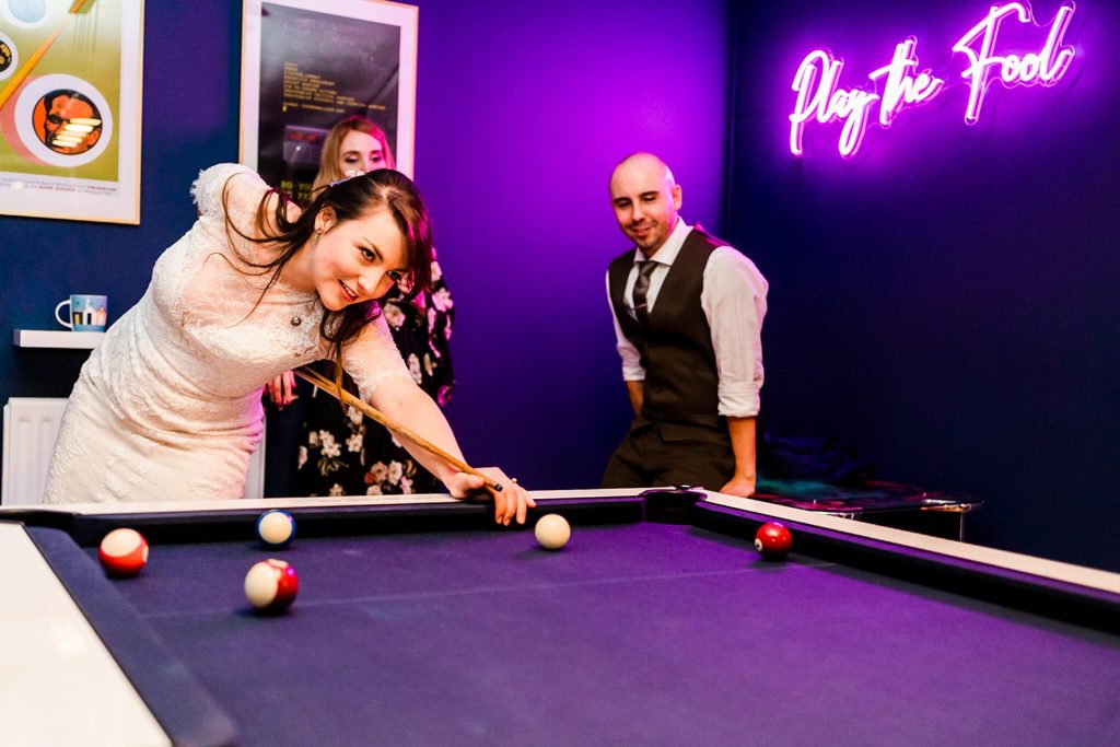 A woman in a white dress plays pool, aiming with a cue stick, while two people watch. A neon sign in the background reads Play the Fool. The room has vibrant blue walls and framed artwork.