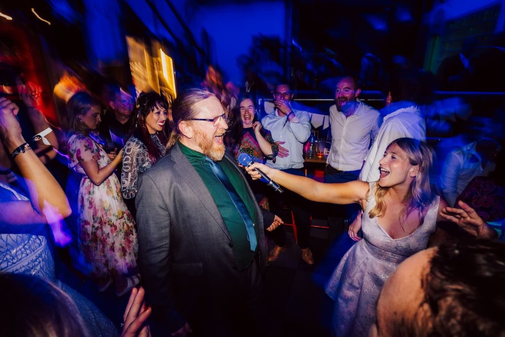 A lively party scene with a bearded man in a suit singing into a microphone. A woman in a light dress holds the mic, smiling. People are dancing and laughing in the colorful, blurred background, creating a fun and energetic atmosphere.