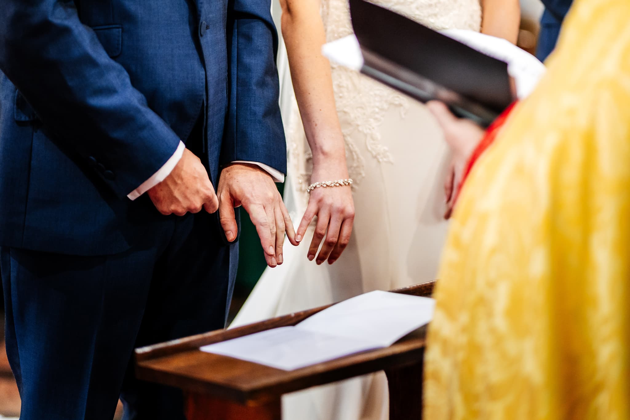 A couple stands side by side, holding hands at a wedding ceremony. The groom wears a blue suit while the bride is in a white gown. They face a person in a yellow robe holding a booklet. A wooden stand with an open book is visible.