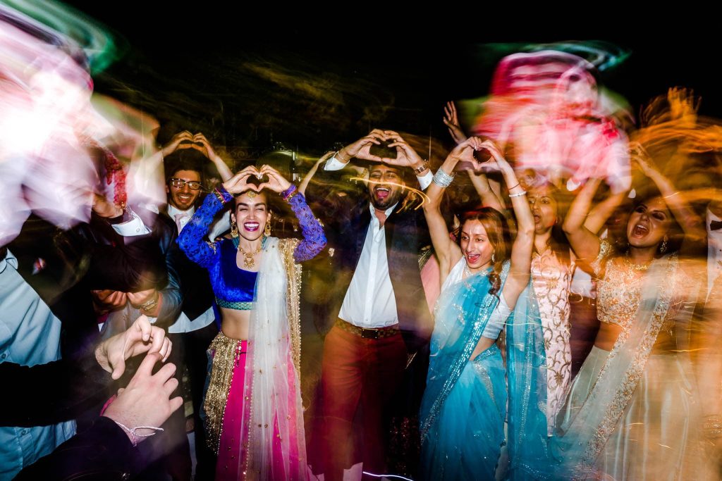 A group of people wearing colorful traditional Indian clothing are dancing energetically in a lively atmosphere. Some form heart shapes with their hands. The background is blurred, giving a dynamic feel to the scene.