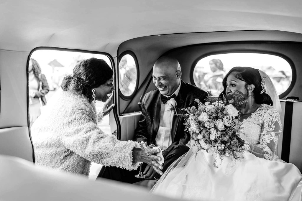 A bride and groom sit in a car, both smiling warmly. The bride holds a bouquet of flowers. A woman, holding the brides hand, leans in through the open car door, sharing in the moment. The scene is captured in black and white.