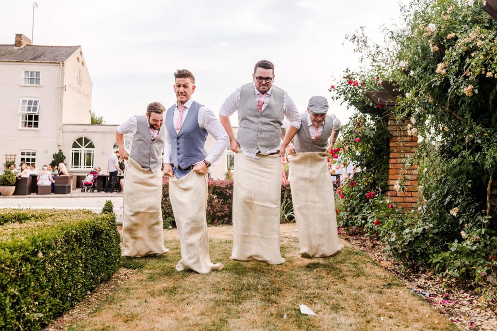 Four men participating in a sack race on a grassy area, dressed in vests and ties, with a garden and building in the background. They are mid-jump, with expressions of focus and amusement.