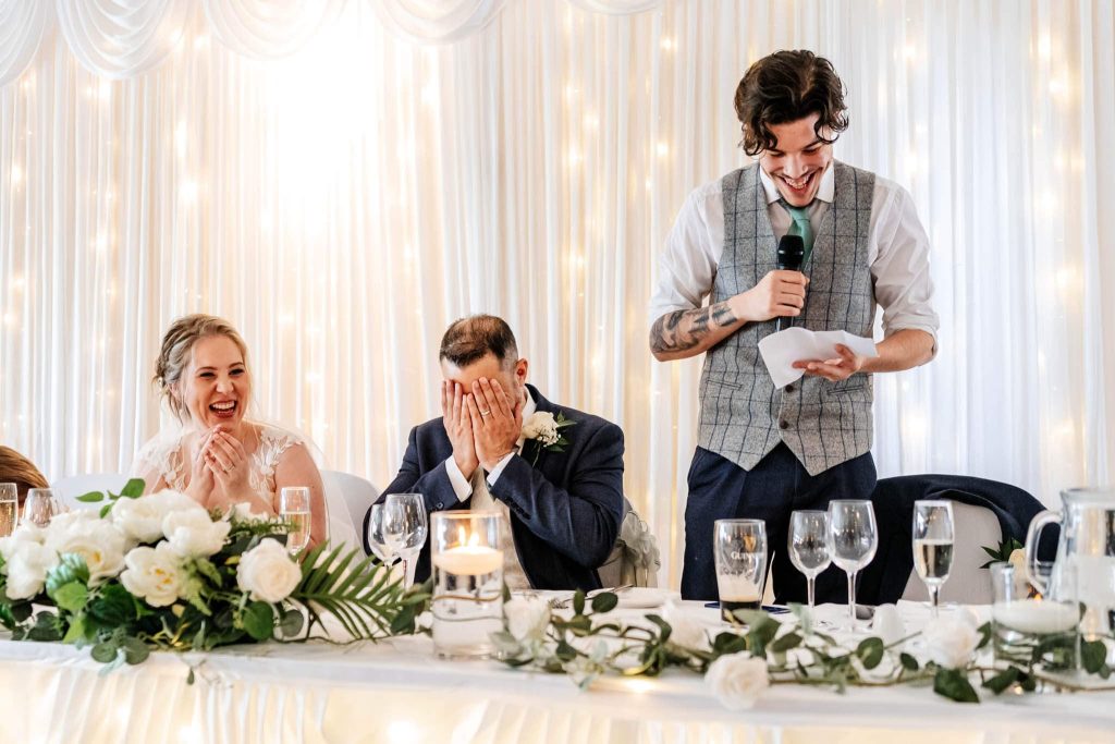 A young man stands at a wedding reception table holding a microphone and speech notes, smiling. A bride laughs, and another man covers his face with his hands. The table is adorned with white roses and candles, with a softly lit backdrop.