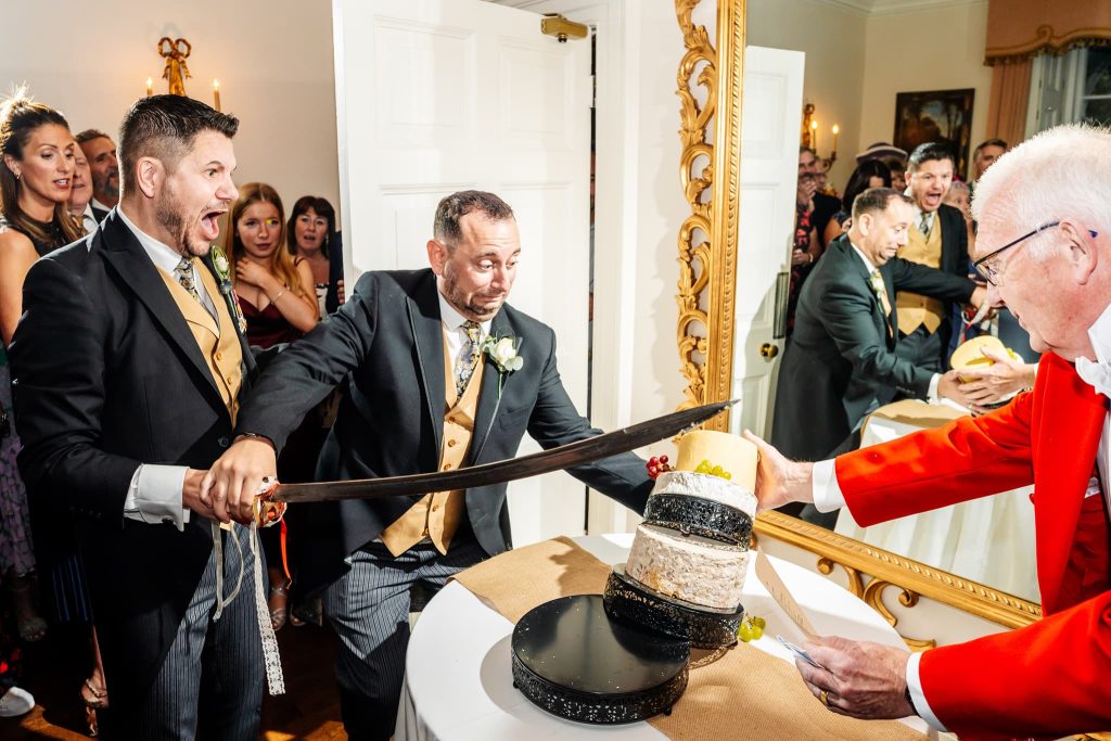 At Sutton Bonington Hall in Nottingham, a man in formal attire playfully cuts a three-tiered cake with a large sword at a wedding reception, assisted by another man. Guests watch and smile in the background as a mirror captures the joyful scene.