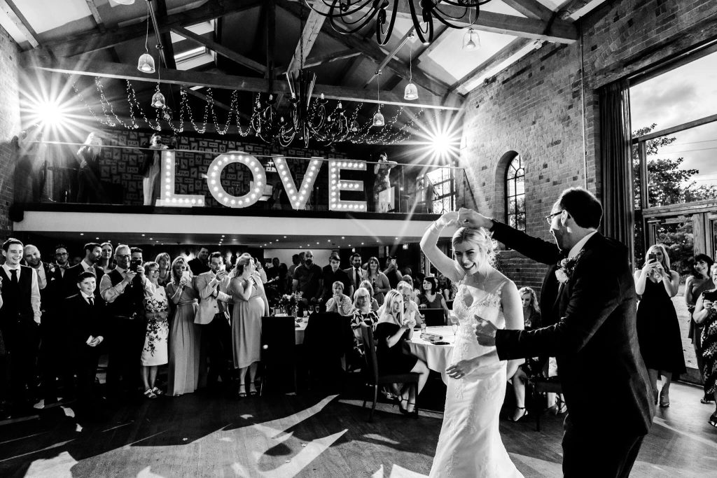 A couple dances at a wedding reception in the grand Carriage Hall, Nottingham, with exposed beams above. Guests surround them, watching and clapping. Behind them, LOVE is illuminated in large letters, as the photographer captures the festive and joyful atmosphere.