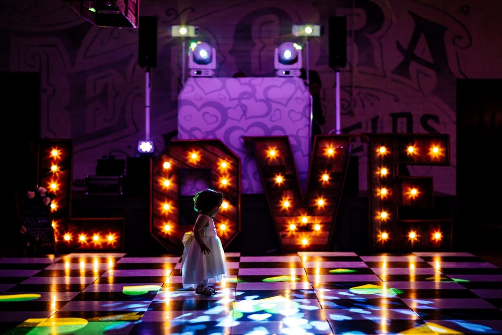 Little flower girl is mesmerised by the disco lights on the dancefloor. She stands in the middle of the dancefloor with lit up lettings spelling LOVE in the background.
