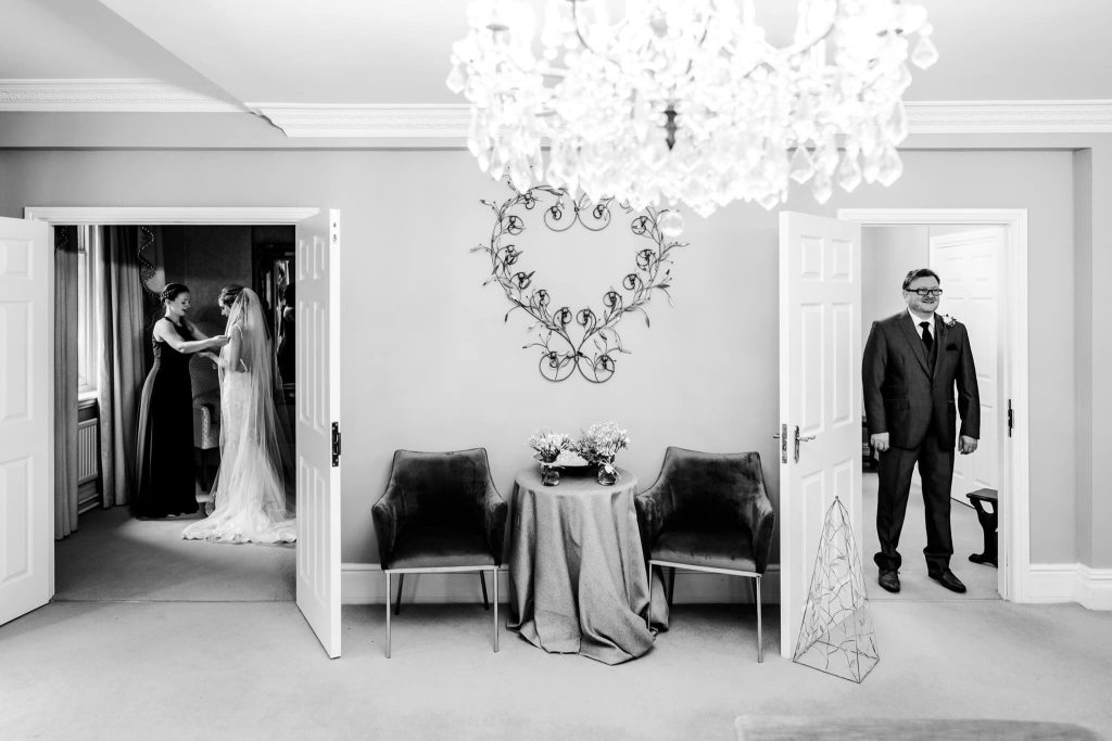 Father of the bride waits outside one door whilst his daughter the bride gets the final adjustments made through another