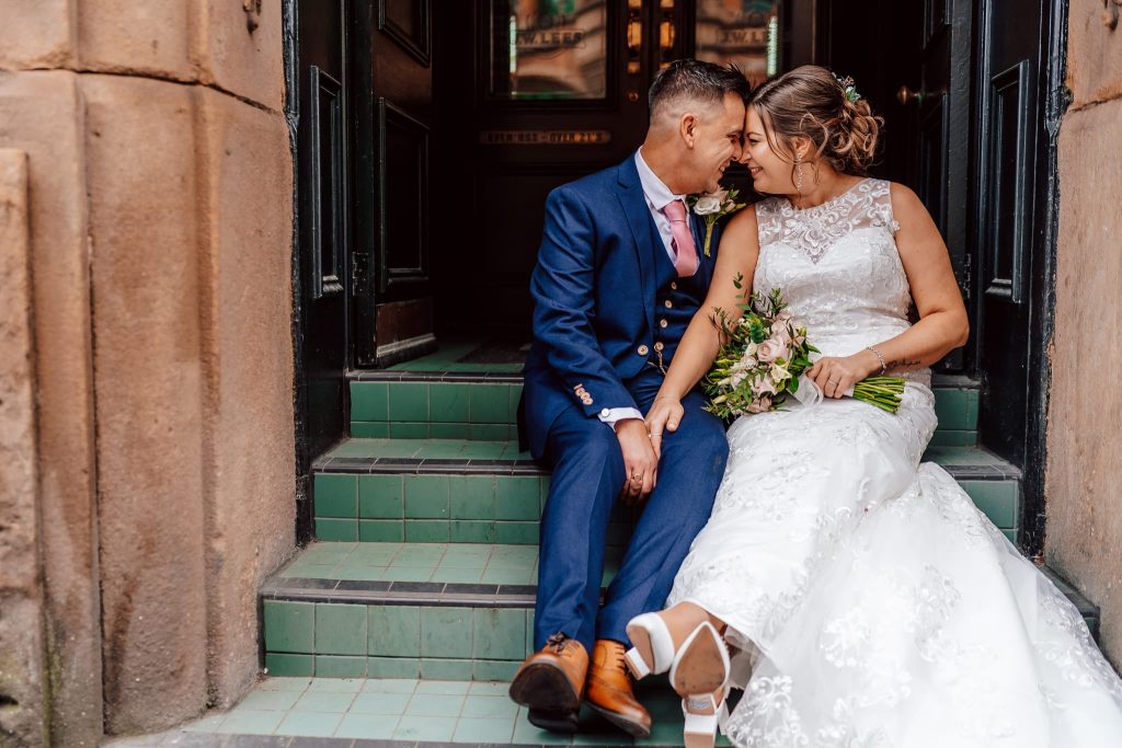 A bride and groom sit closely on the green-tiled steps of a charming Manchester wedding venue Rain Bar, smiling at each other. She holds a floral bouquet next to her wedding dress, while he wears a navy suit with a pink tie.