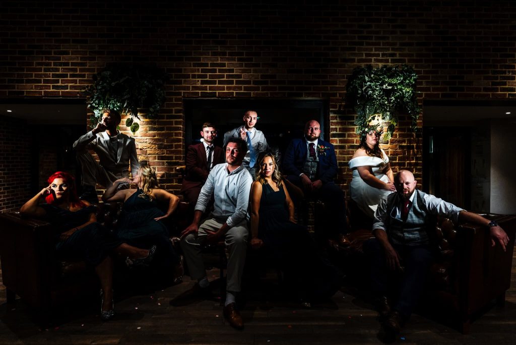 A group of people in formal attire pose dramatically in low lighting against a brick wall backdrop at Foxtail Barns Staffordshire. Some are sitting, while others stand, creating a theatrical and moody atmosphere reminiscent of alternative wedding photography.