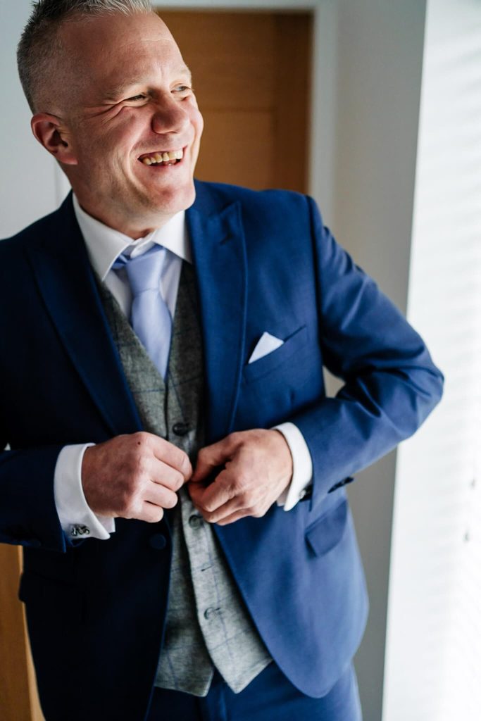 A man in a blue suit and gray waistcoat is smiling while buttoning his jacket. He stands near a window with blinds, allowing soft light to illuminate his face, capturing the essence of relaxed wedding photography.