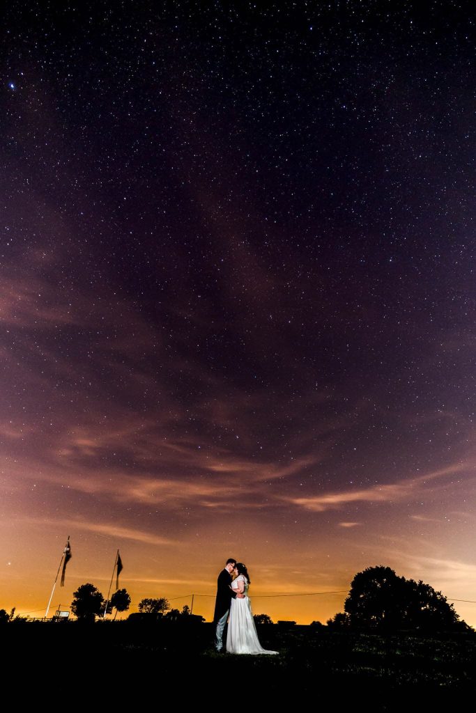 A couple embraces under a vast, starry night sky, capturing the essence of night time wedding photos. The horizon glows with a warm orange hue, silhouetting trees and flags. Wispy clouds are scattered amidst the stars, creating a romantic and serene atmosphere.