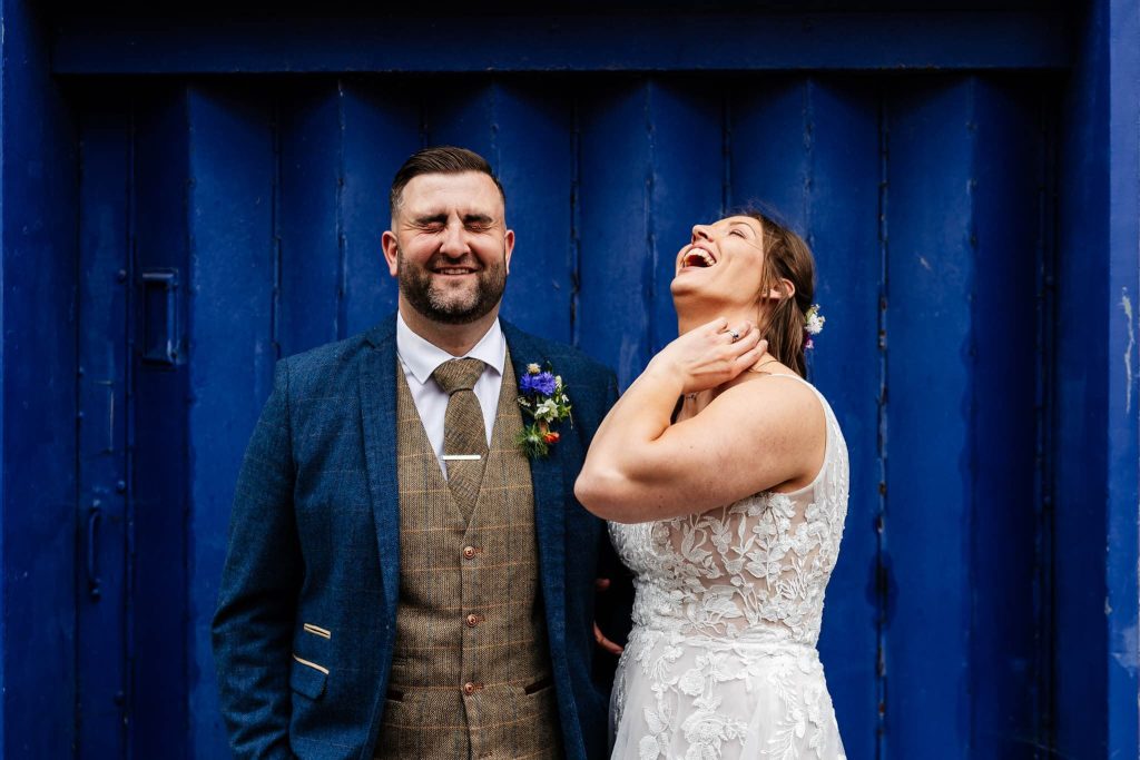 A joyful bride and groom stand in front of a blue door at Westgates Suites Long Eaton. The groom wears a blue suit with a brown vest, and the bride wears a white lacy gown. Both are laughing, creating a cheerful atmosphere.