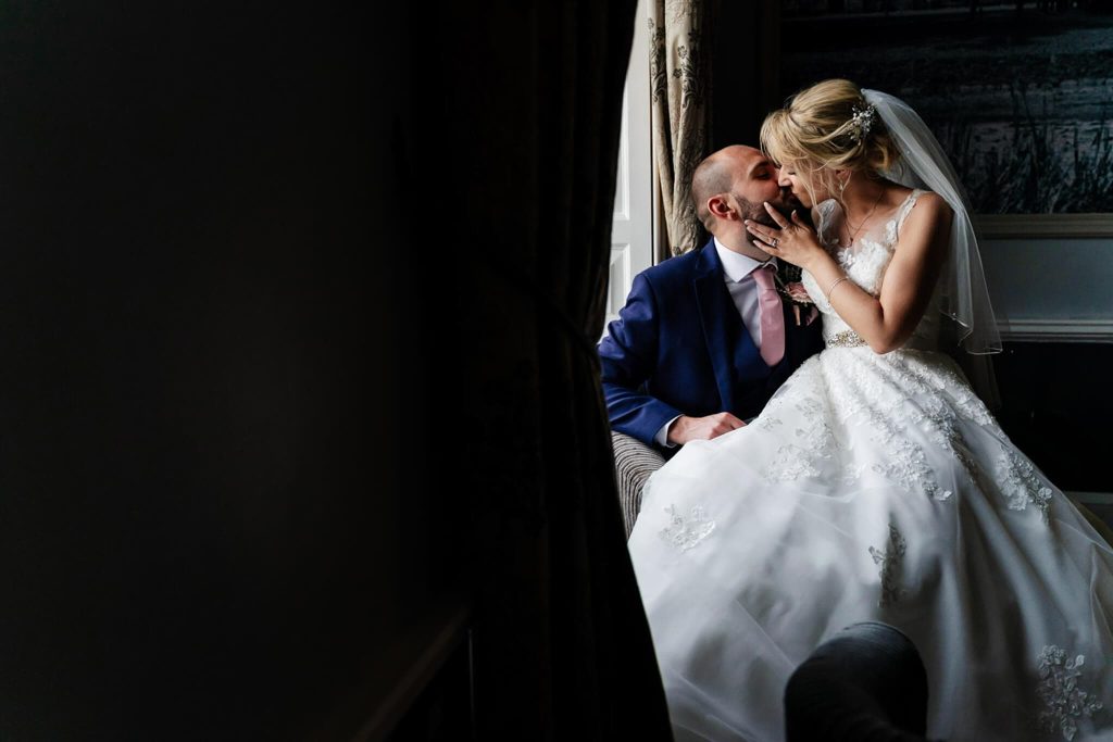 A bride and groom share a kiss on a windowsill at Colwick Hall Nottingham. The bride wears a flowing white gown and veil, while the groom dons a dark blue suit with a pink tie. Soft lighting filters through the curtains, casting an enchanting glow on their intimate moment.