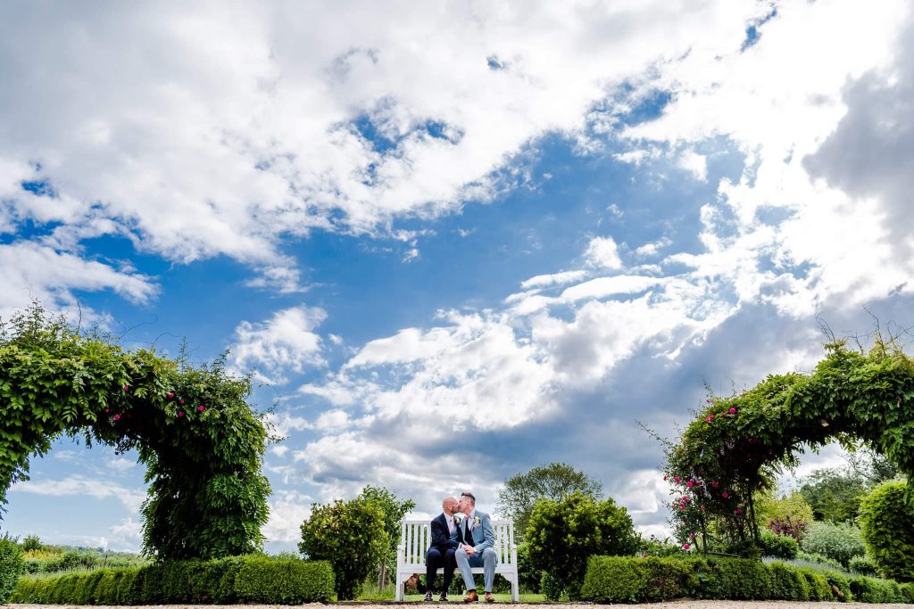 A couple sits on a white bench in a lush garden at Stapleford Park Hotel, framed by two green archways and vibrant bushes. The sky, filled with dramatic clouds, adds depth to the serene setting, perfect for LGBTQ+ weddings.