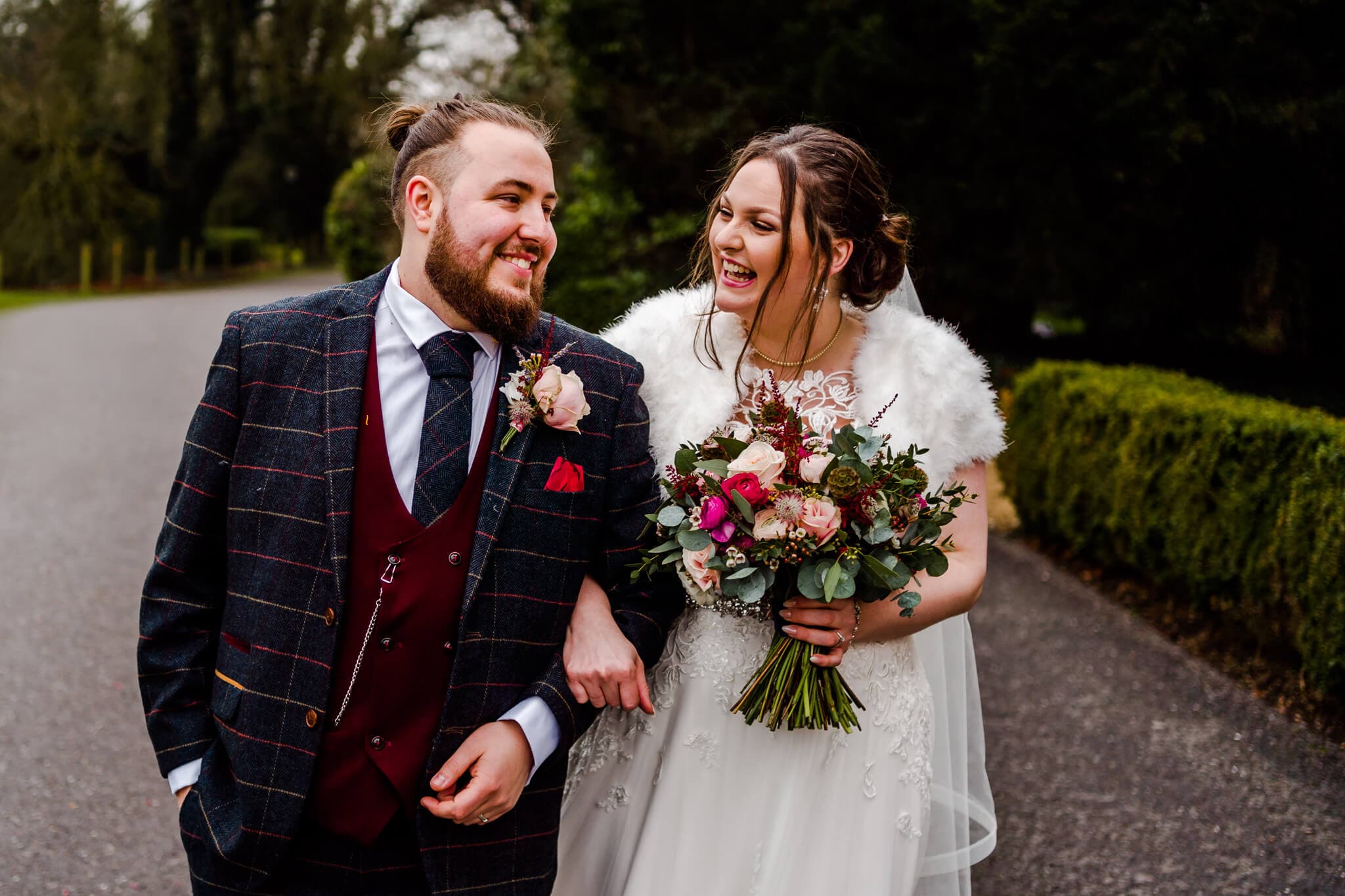 A joyful couple walks down a path at Ansty Hall, arm in arm, dressed in wedding attire. The groom dons a dark plaid suit with a burgundy vest, while the bride, in a white gown with a fur wrap, holds a colourful bouquet and smiles warmly at their winter wedding.