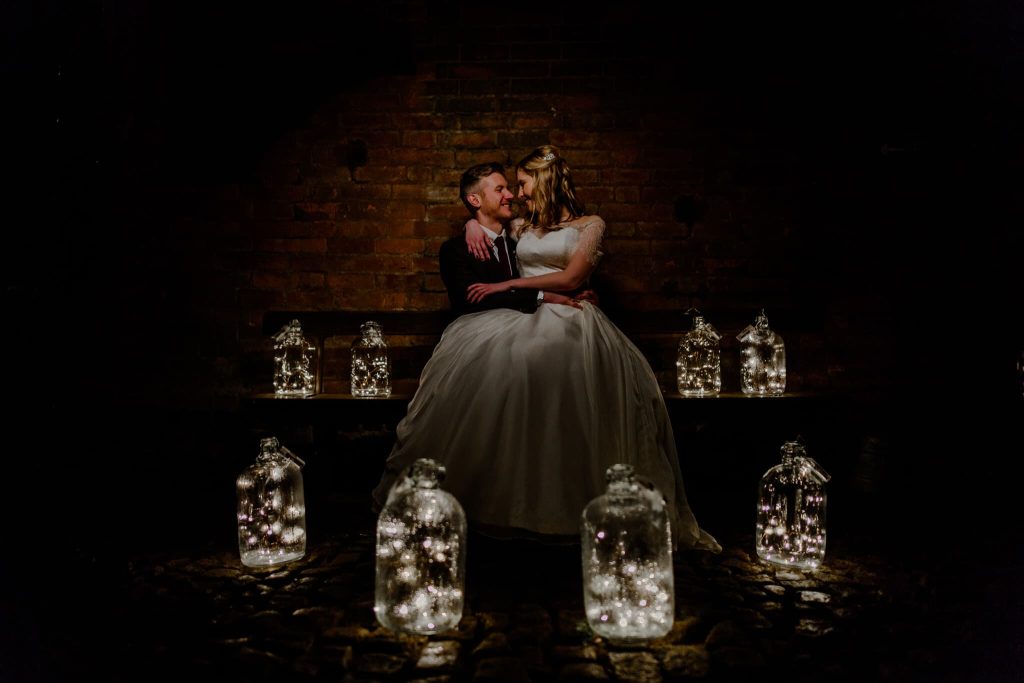 A bride and groom sit closely on a bench surrounded by glowing fairy lights inside glass jars. The backdrop of a dimly lit brick wall enhances the warm, intimate atmosphere. These creative wedding photos capture the couple smiling warmly at each other, reflecting their joy and love.