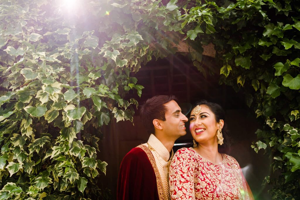 A couple in traditional attire stands closely under a lush archway at Keythorpe Manor, surrounded by vibrant greenery. The sunlight casts a soft glow around them, capturing the essence of Asian wedding photography. The woman smiles brightly with ornate jewelry, as the man gazes at her affectionately.