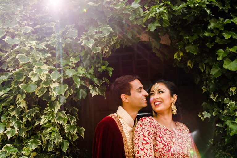 A couple in traditional attire stands closely under a lush archway at Keythorpe Manor, surrounded by vibrant greenery. The sunlight casts a soft glow around them, capturing the essence of Asian wedding photography. The woman smiles brightly with ornate jewelry, as the man gazes at her affectionately.