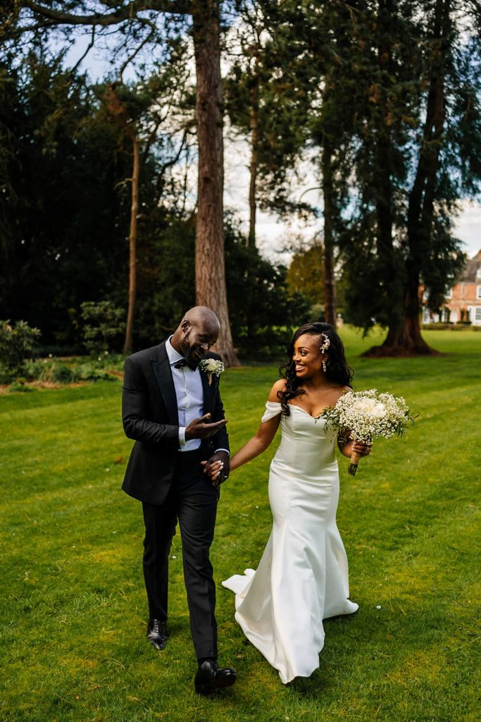 A couple walks hand in hand on a grassy field at Sutton Bonington Hall, surrounded by tall trees. The bride wears a white gown and holds a bouquet, while the groom is in a black suit, adjusting his boutonniere. They share a joyful, intimate moment outdoors in Nottinghams picturesque setting.