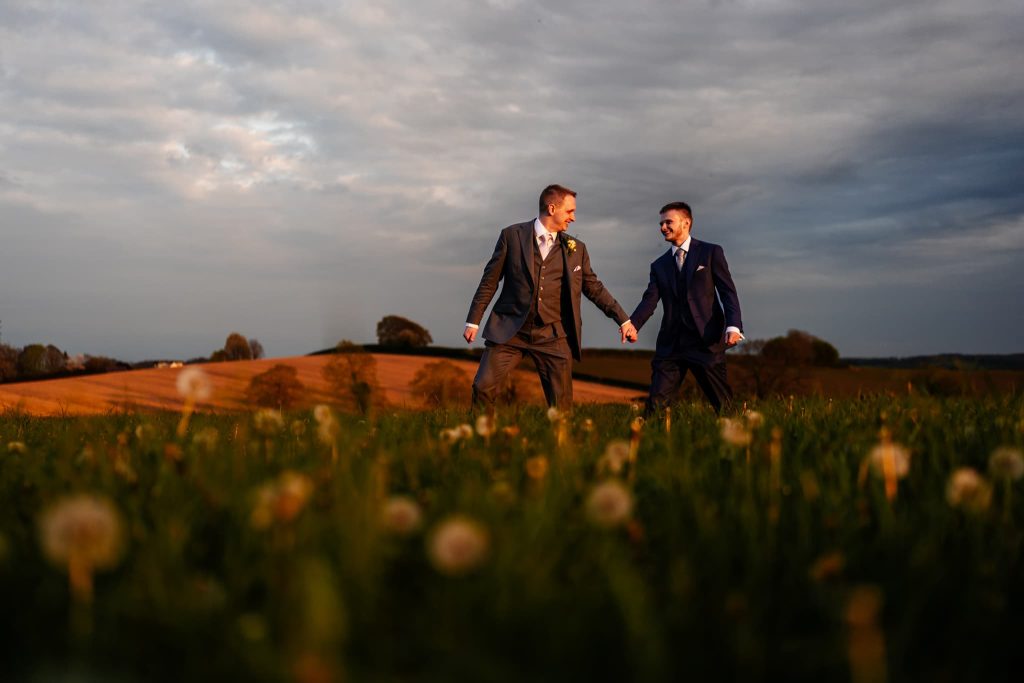 Two men in suits hold hands and walk through a field of dandelions near Donington Park Farmhouse, under a partly cloudy sky. The landscape features rolling hills and a warm sunset, creating a romantic atmosphere in Leicestershire.