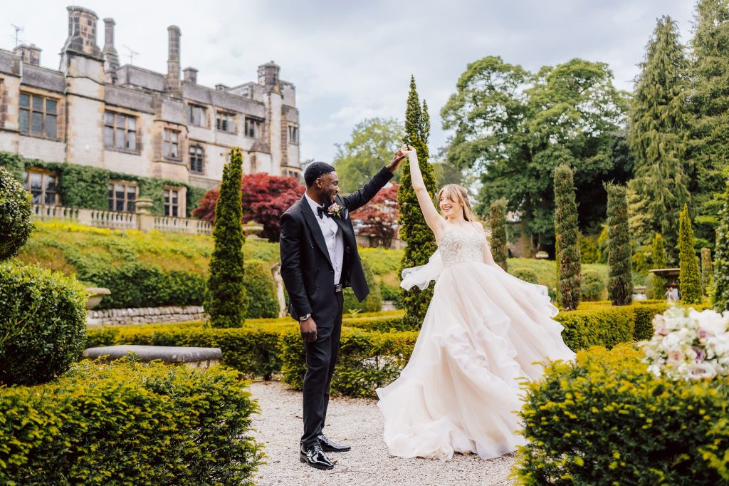 A bride in a flowing gown and a groom in a suit dance elegantly at Thornbridge Estate. The backdrop features a historic stone building, green hedges, and tall trees, creating a romantic atmosphere perfect for their Nigerian fusion wedding.